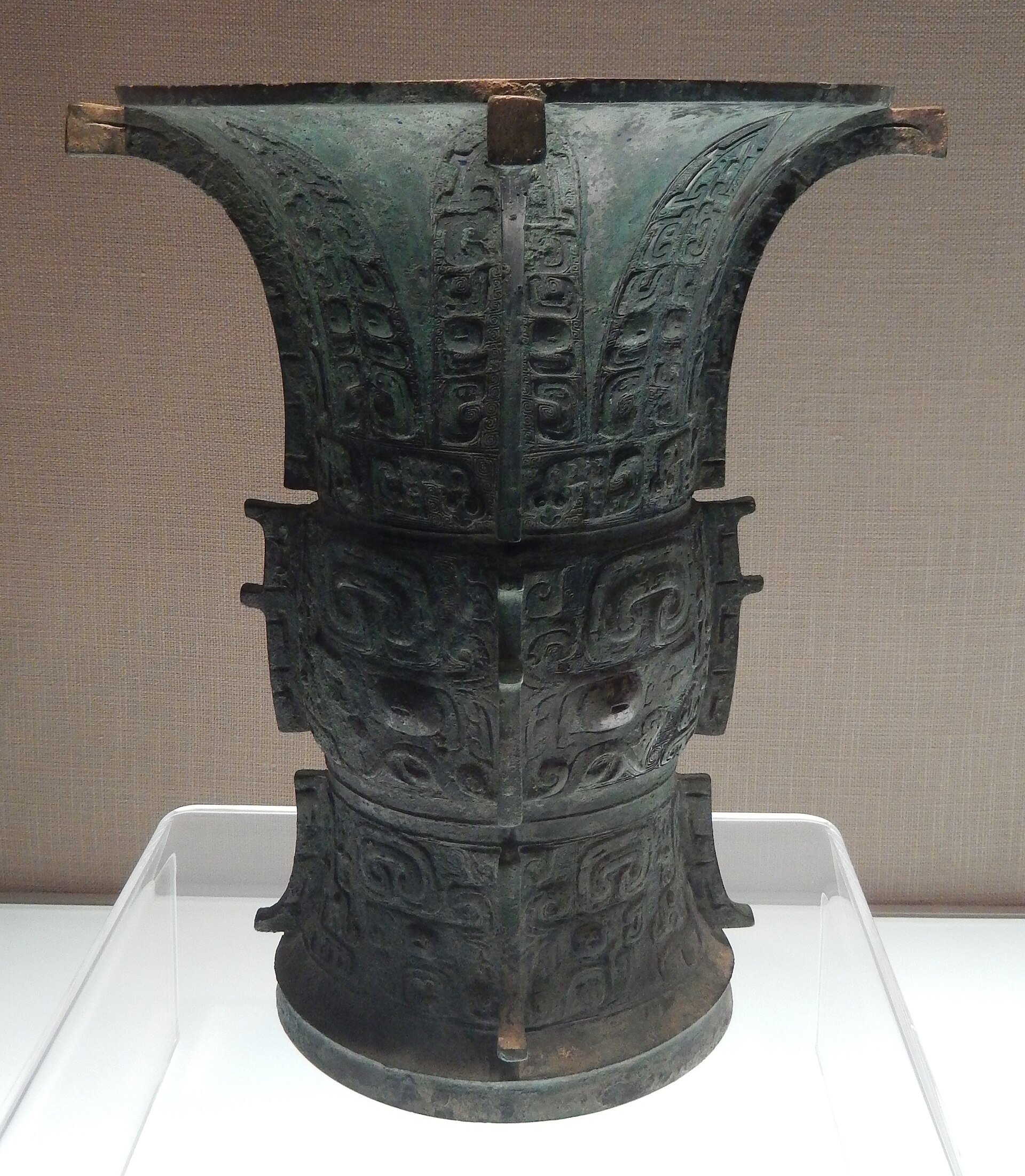 A Western Zhou bronze zun wine vessel with a flared mouth and flanged sides, covered in relief decoration, on a white plinth