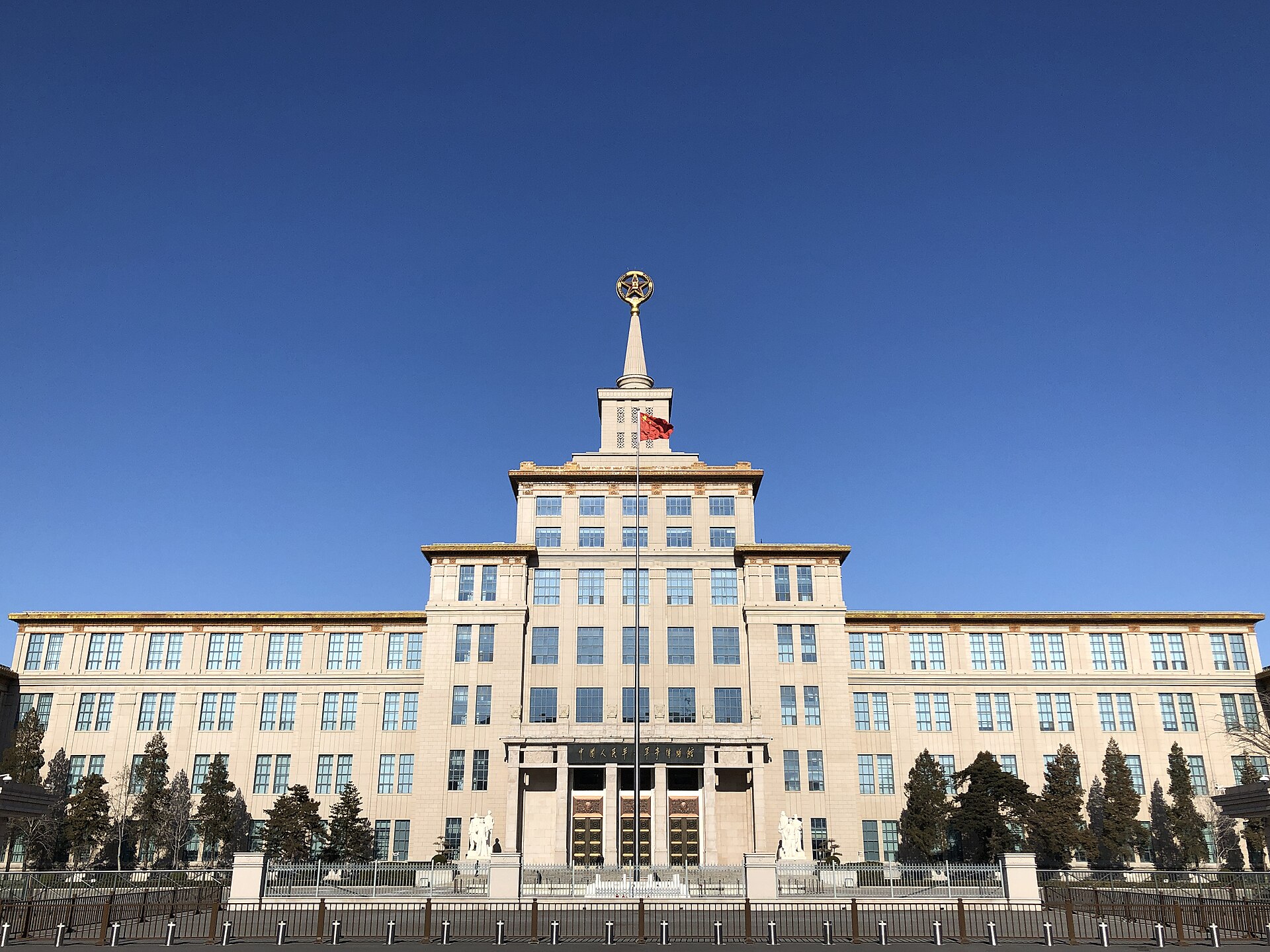 The cream stone facade of the Military Museum of the Chinese People's Revolution in Beijing, with a central tower topped by a gilded star and a red flag in front