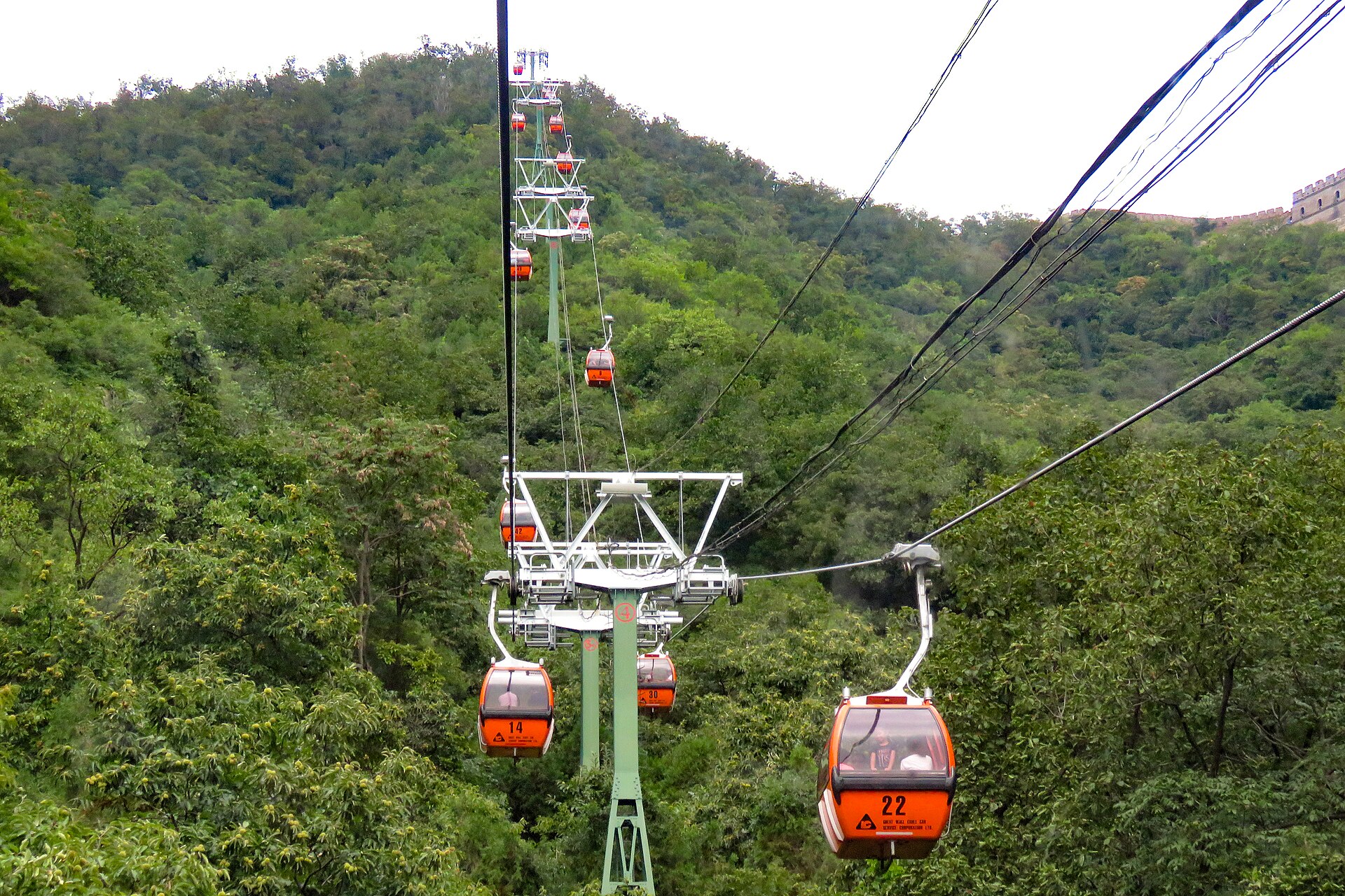 Orange gondolas of the Mutianyu cable car climbing a forested hillside towards the Great Wall, with a watchtower visible at the top right