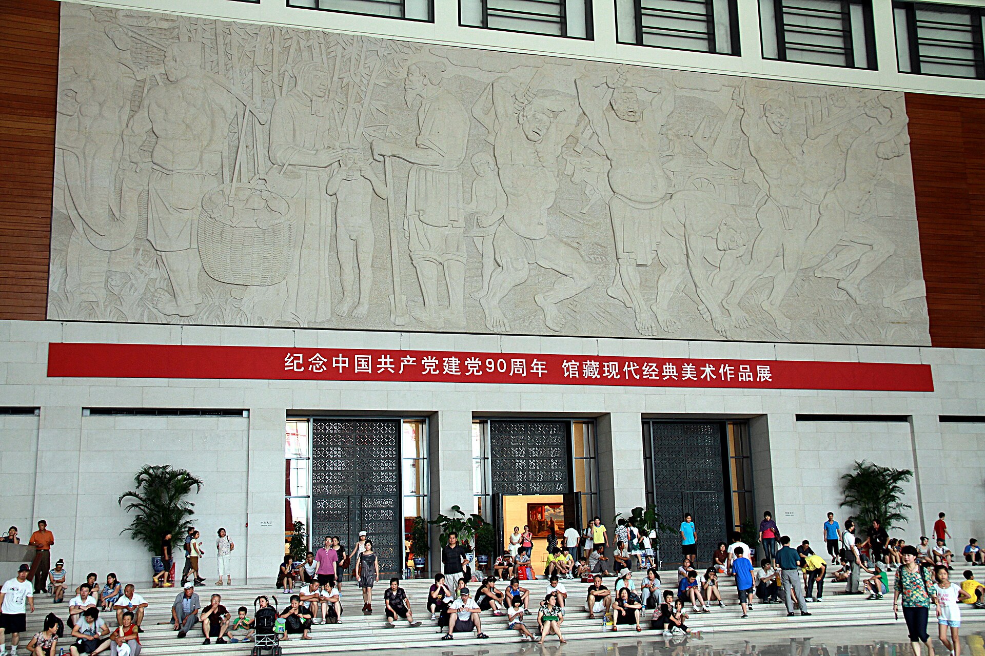 Visitors sitting on the steps of the National Museum of China's central hall beneath a large stone relief and a red exhibition banner