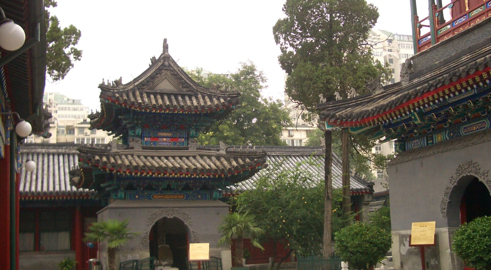 Painted pavilions and a two-storey tower surround a paved courtyard at Niujie Mosque in Beijing, with trees and potted plants.