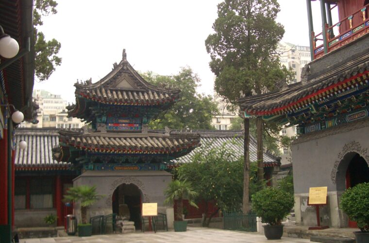 Painted pavilions and a two-storey tower surround a paved courtyard at Niujie Mosque in Beijing, with trees and potted plants.