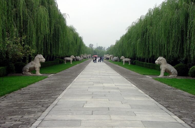 Paired stone lions and other animals line the willow-bordered Sacred Way at Beijing's Ming Tombs, with visitors in the distance.