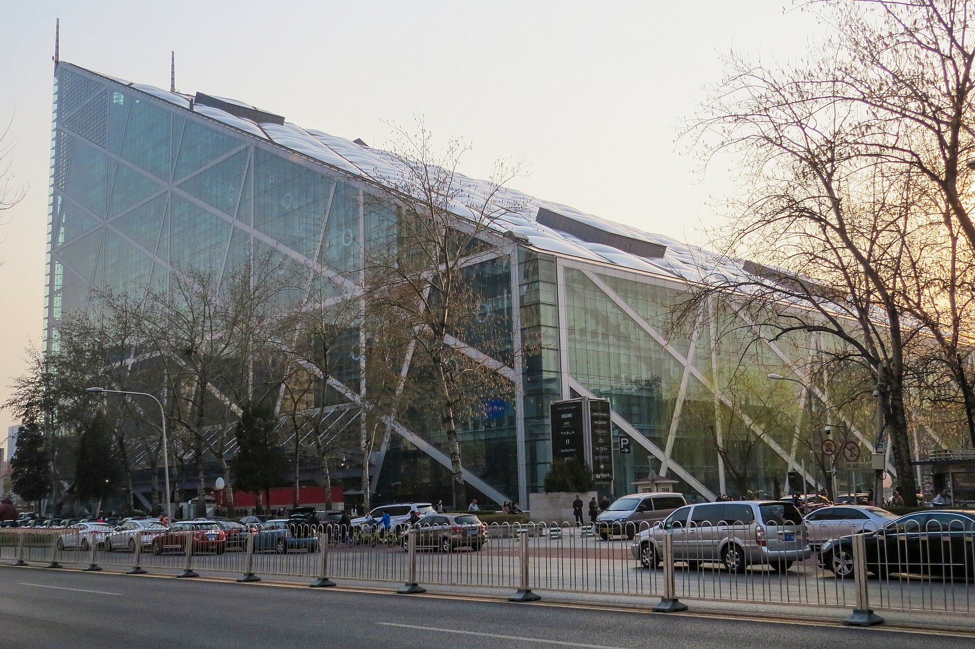 Parkview Green's sloping glass exterior and diagonal steel braces rise behind roadside trees and traffic in Beijing.