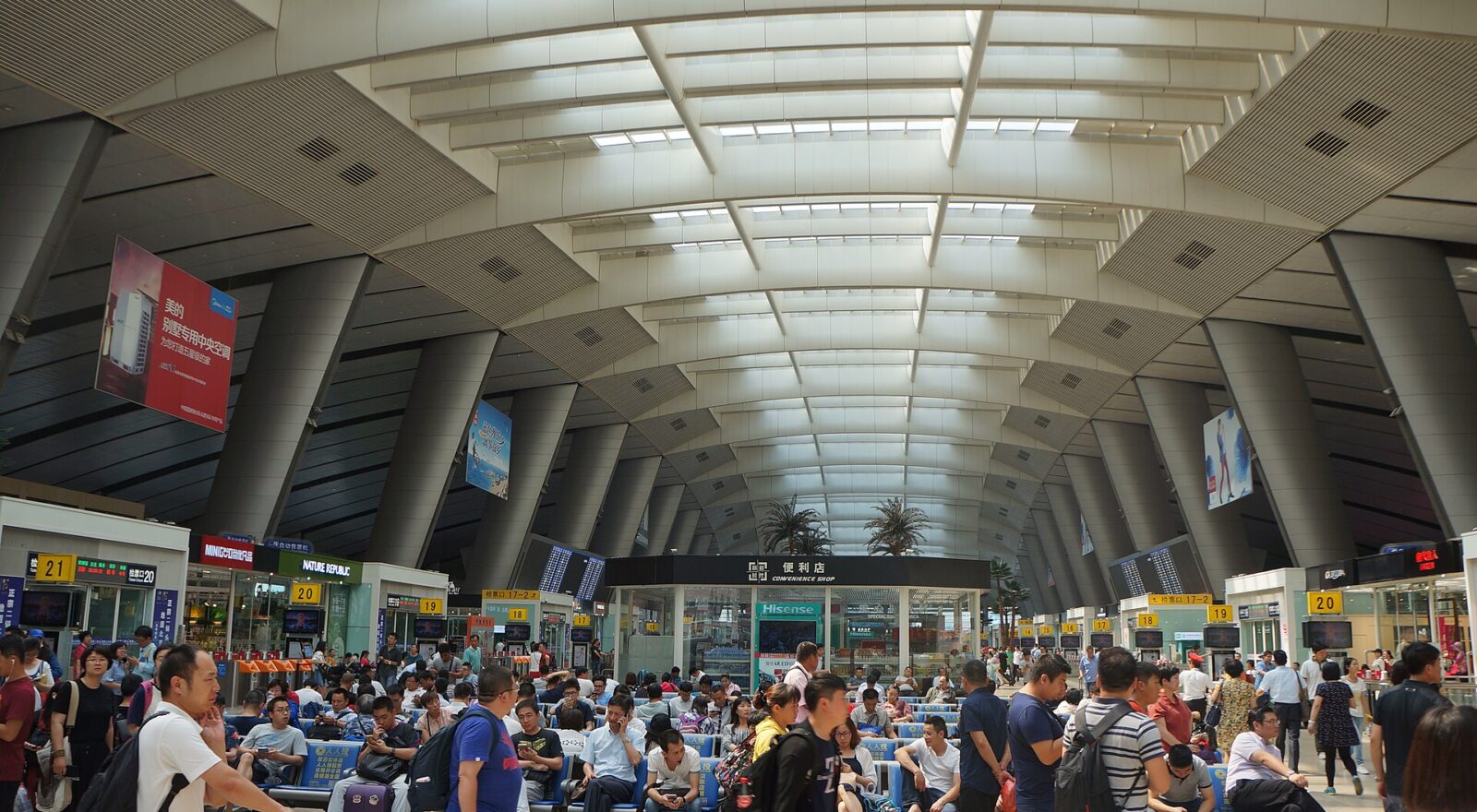 Passengers sit and stand with luggage beneath the high skylit roof and numbered boarding gates at Beijing South Railway Station.