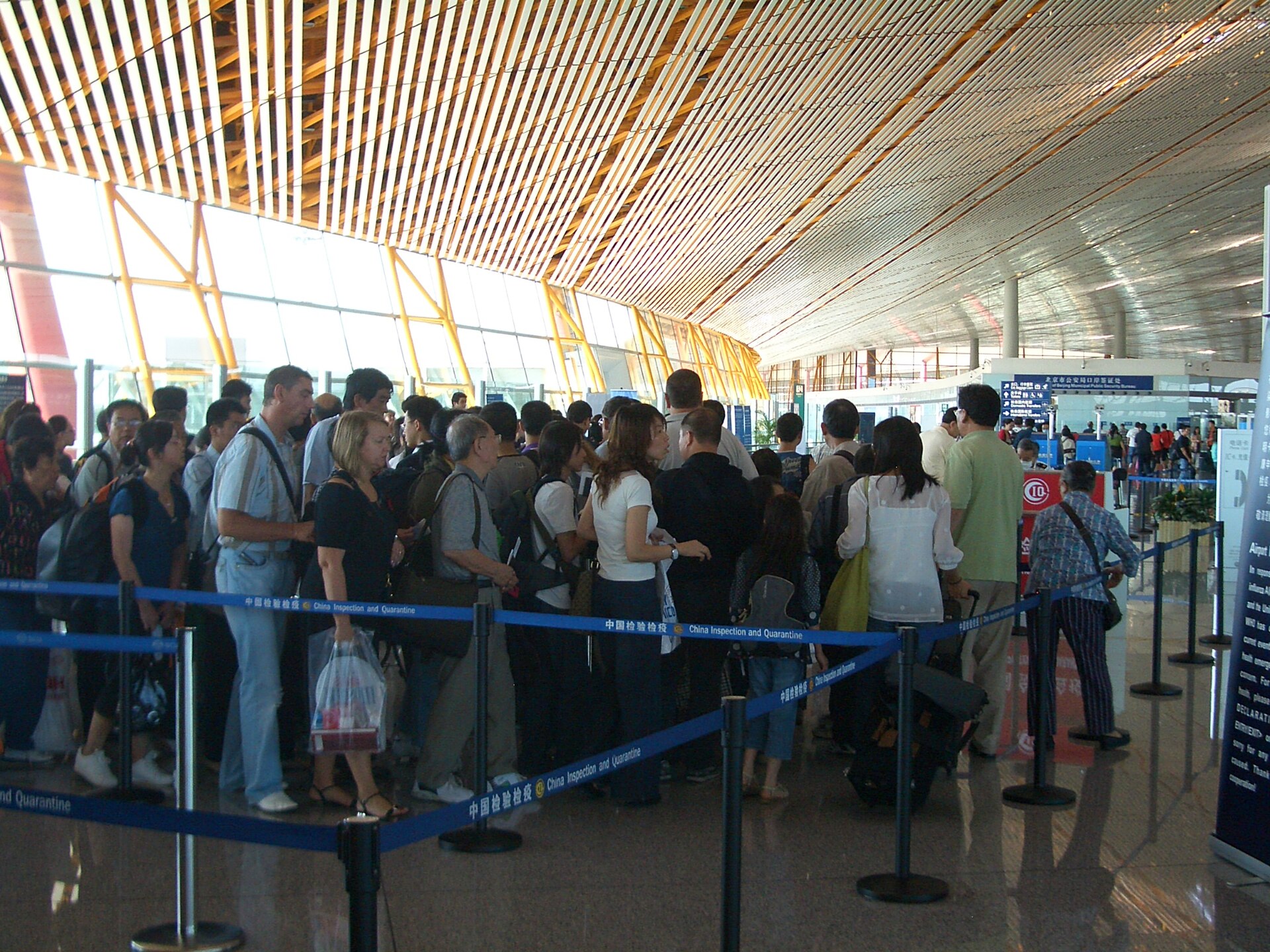Passengers wait between blue queue barriers beneath the high slatted ceiling of Beijing Capital Airport Terminal 3 immigration hall.
