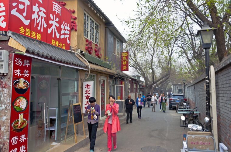 Pedestrians pass a small noodle restaurant with red signs on tree-lined Fangzhuanchang Hutong in Beijing.