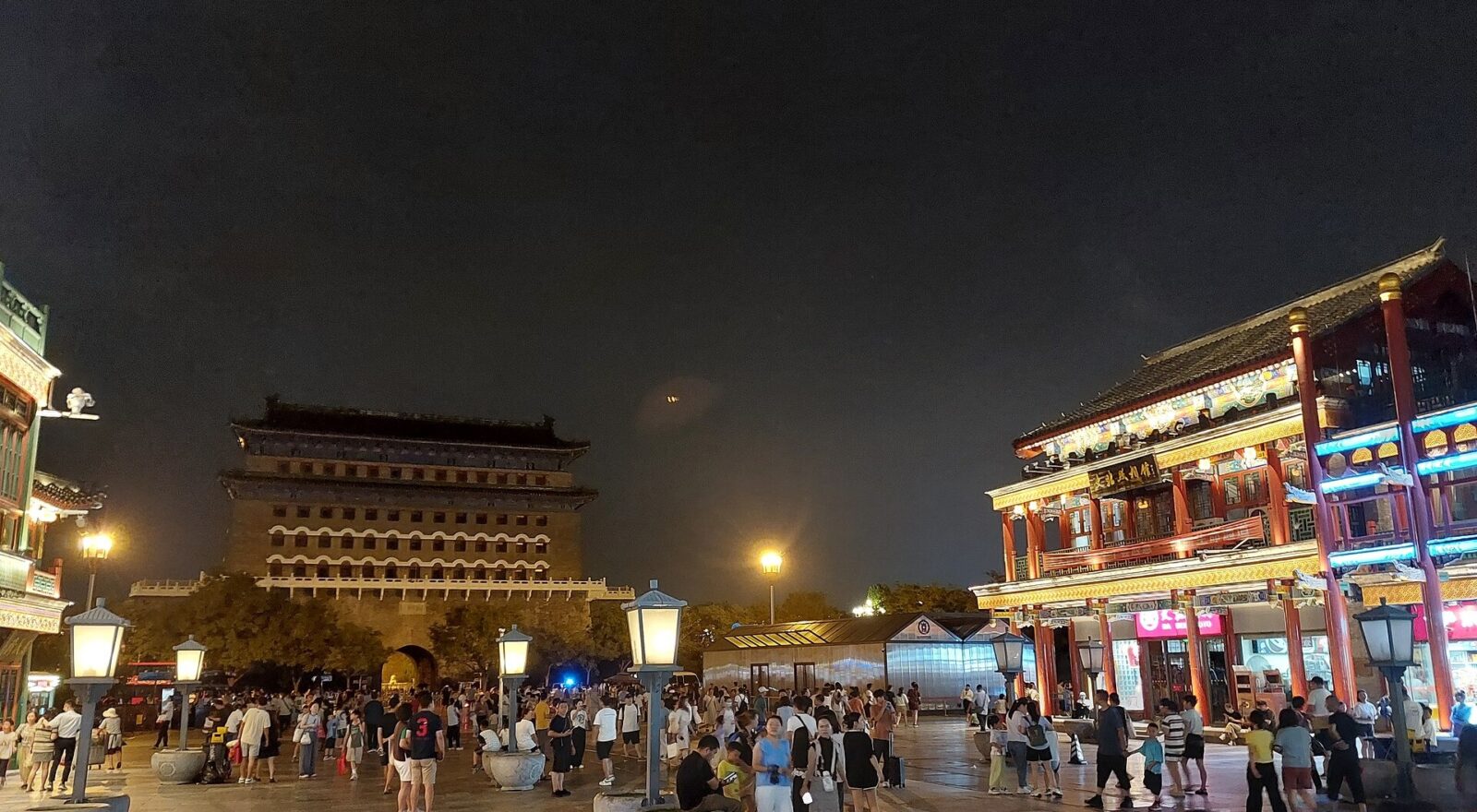 Pedestrians walk along illuminated Qianmen Dajie in Beijing at night, with the archery tower rising beyond the shops.