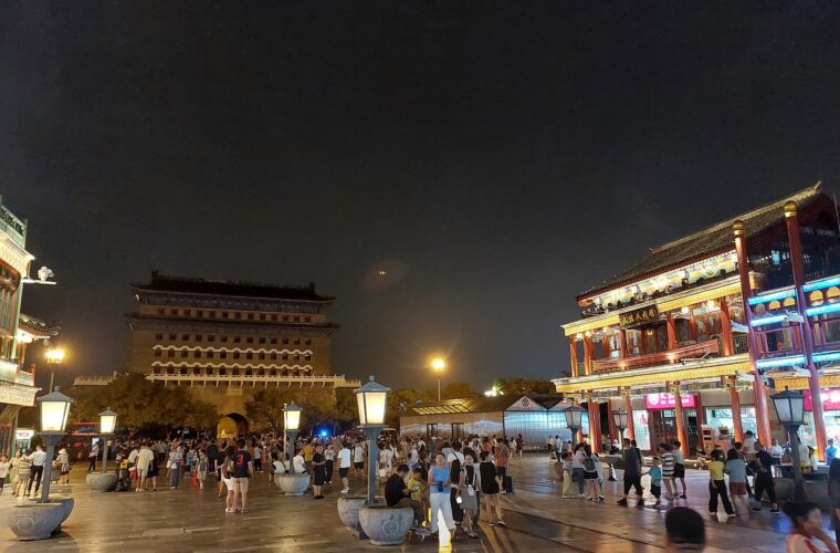 Pedestrians walk along illuminated Qianmen Dajie in Beijing at night, with the archery tower rising beyond the shops.