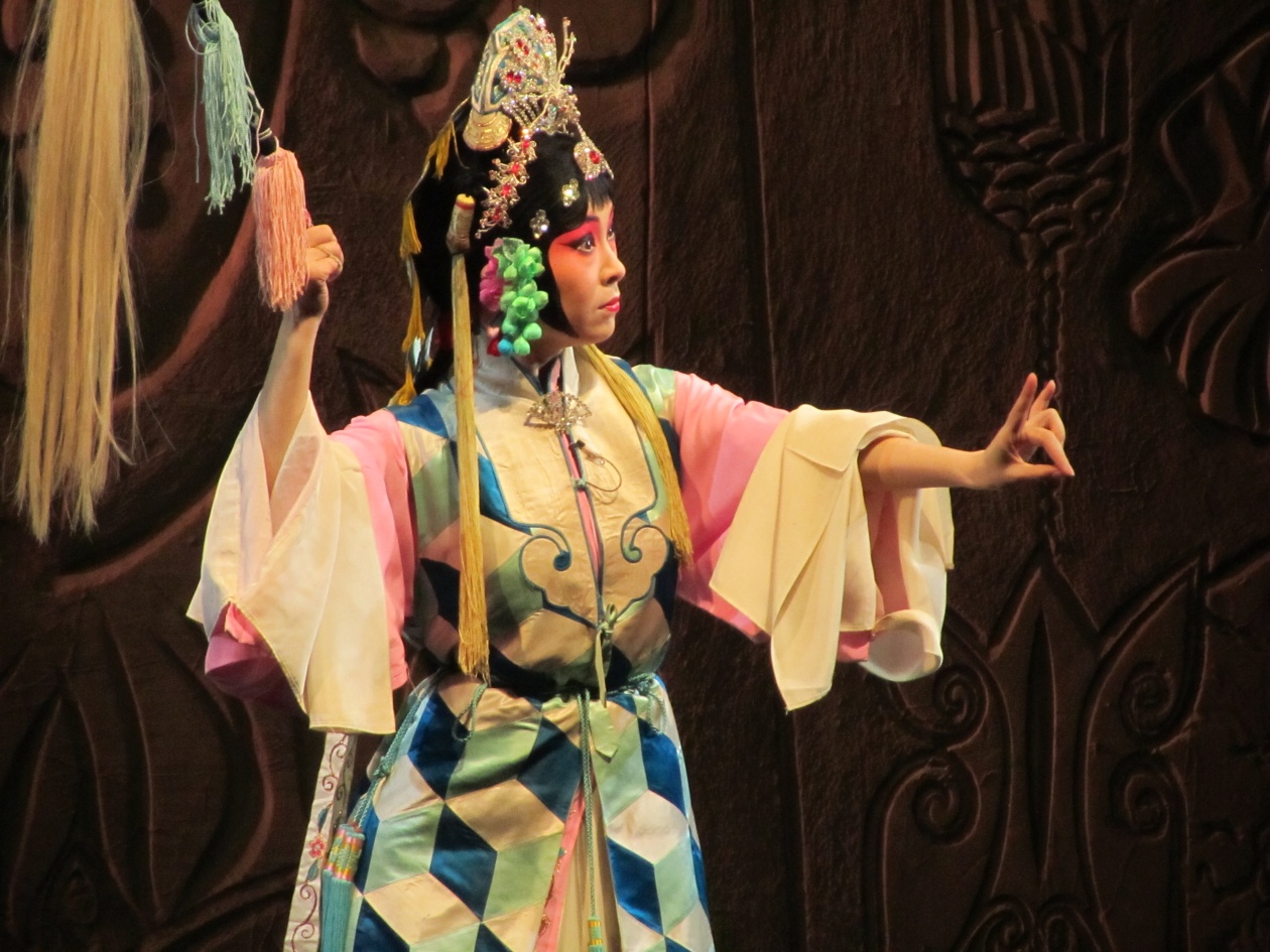 A Peking Opera performer in Beijing extends a hand while holding a tasselled prop, wearing flowing sleeves and an ornate headdress.