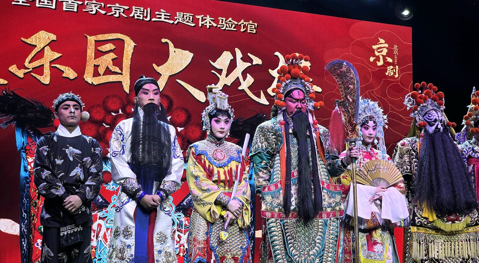 Peking Opera performers in Beijing wear embroidered robes, ornate headdresses and painted faces against a red stage backdrop.