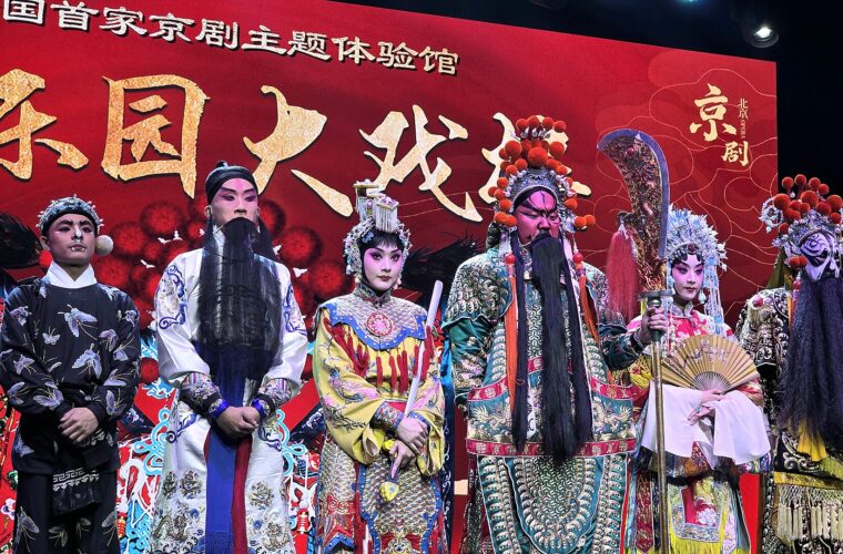 Peking Opera performers in Beijing wear embroidered robes, ornate headdresses and painted faces against a red stage backdrop.