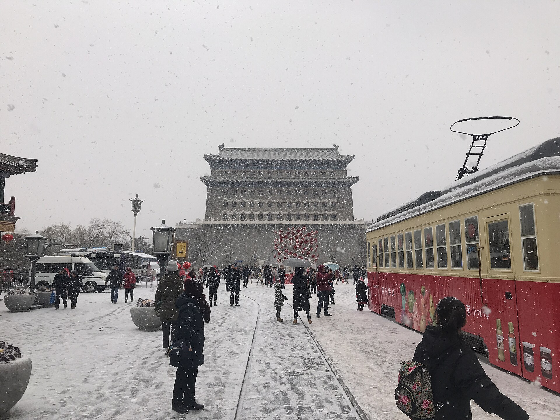 People in winter coats walk through falling snow on Beijing's Qianmen Street beside a red-and-yellow tram and the archery tower.