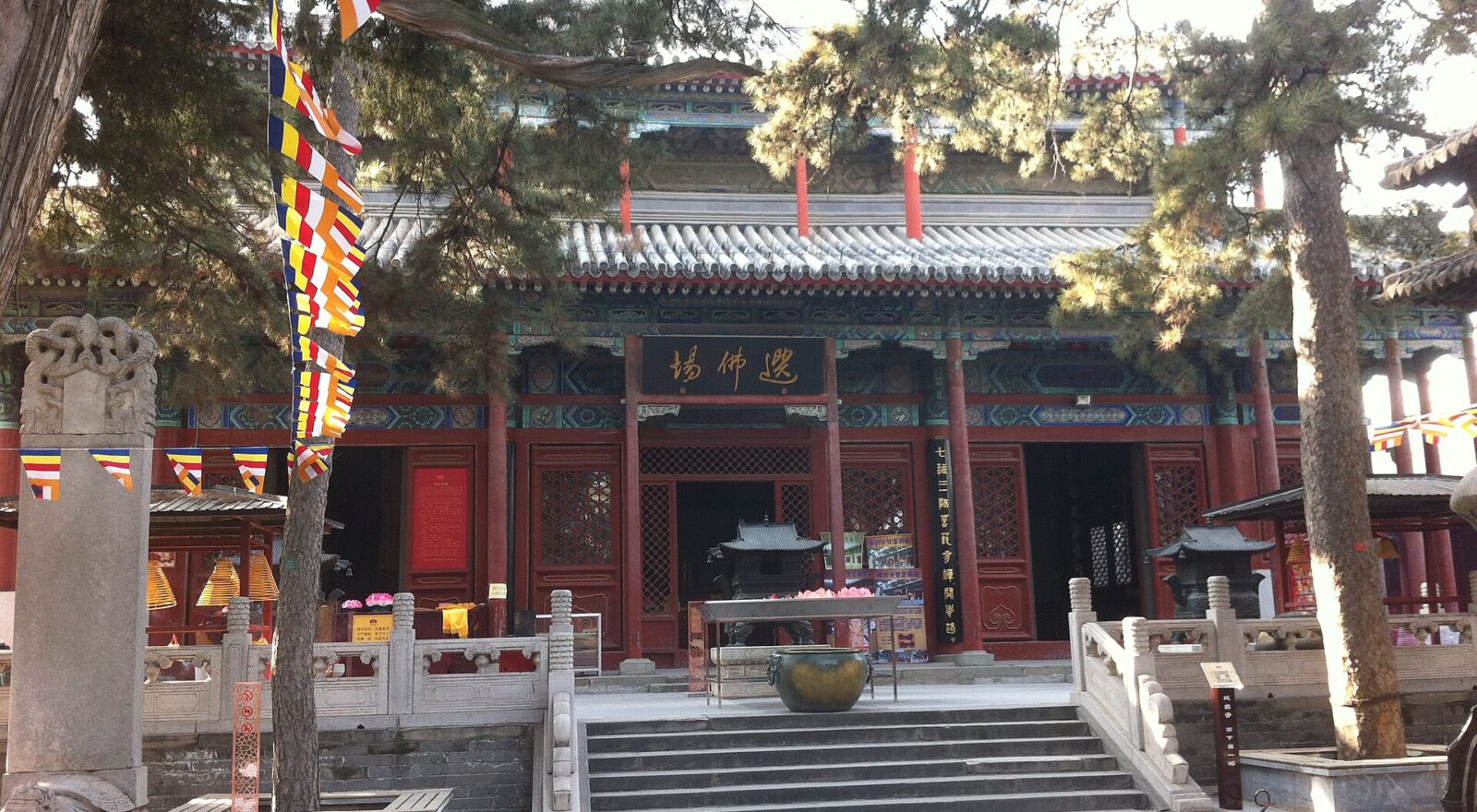 Pine branches and Buddhist flags frame a red hall and stone stairway at Jietai Temple in Beijing.