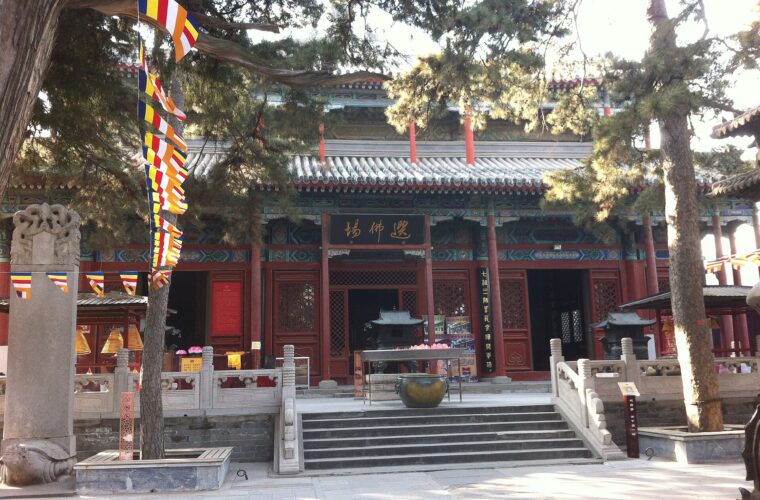 Pine branches and Buddhist flags frame a red hall and stone stairway at Jietai Temple in Beijing.