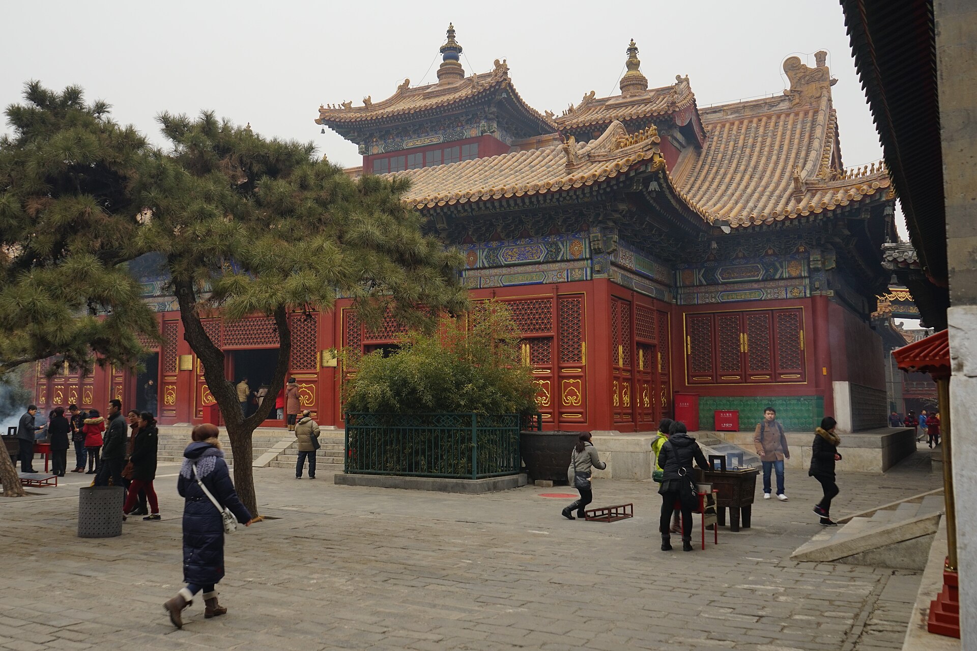 A pine tree shades visitors and incense burners beside a red hall with yellow tiled roofs at Beijing's Lama Temple.