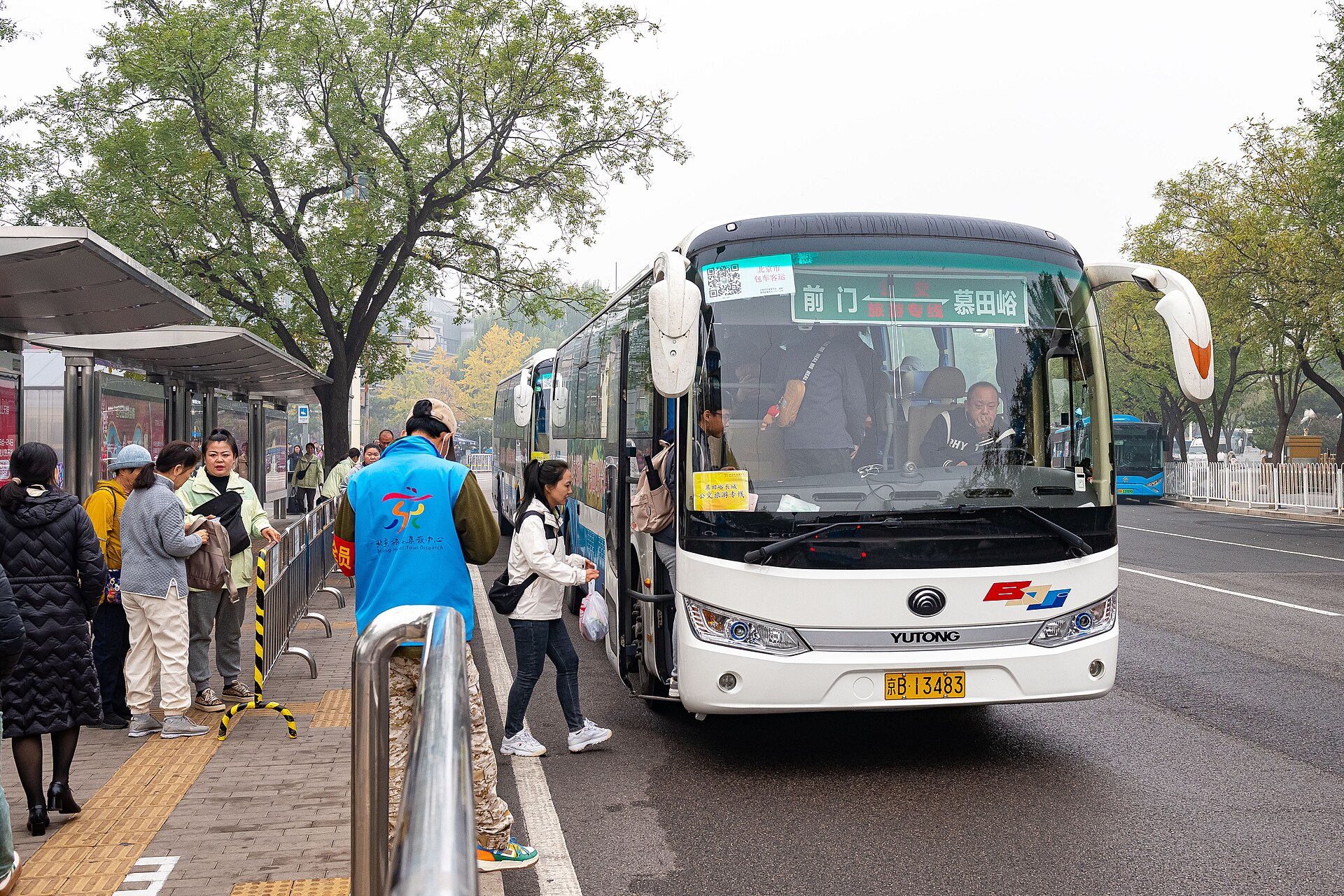 Passengers boarding a white coach at a roadside stop in Beijing, its destination board reading Qianmen to Mutianyu, with a staff member in a blue vest beside the queue