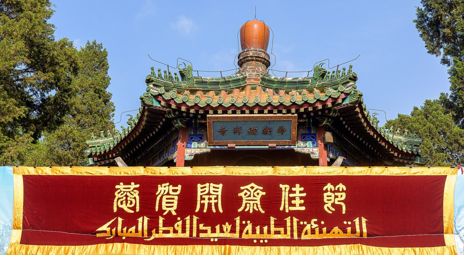 A red Arabic and Chinese banner hangs beneath Niujie Mosque's green tiled entrance roof against a clear Beijing sky.
