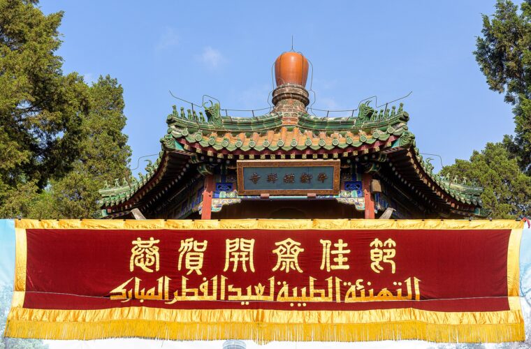A red Arabic and Chinese banner hangs beneath Niujie Mosque's green tiled entrance roof against a clear Beijing sky.