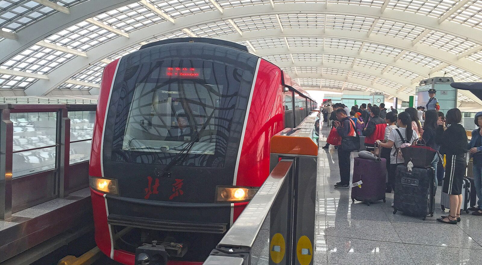 A red Capital Airport Express train arrives at Beijing Capital Airport Terminal 3 beside passengers waiting with wheeled luggage.