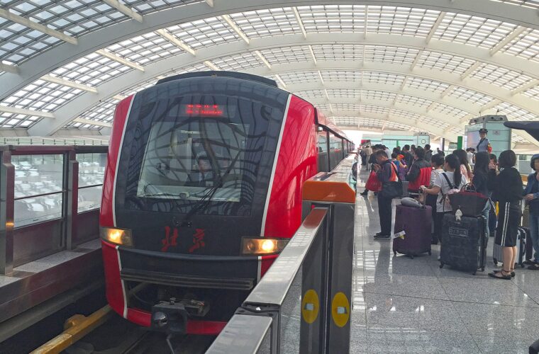 A red Capital Airport Express train arrives at Beijing Capital Airport Terminal 3 beside passengers waiting with wheeled luggage.