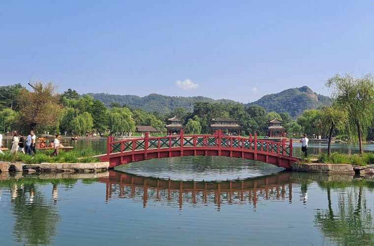 A red footbridge spans the lake at Chengde Mountain Resort, with pavilions, willows and wooded hills beyond.