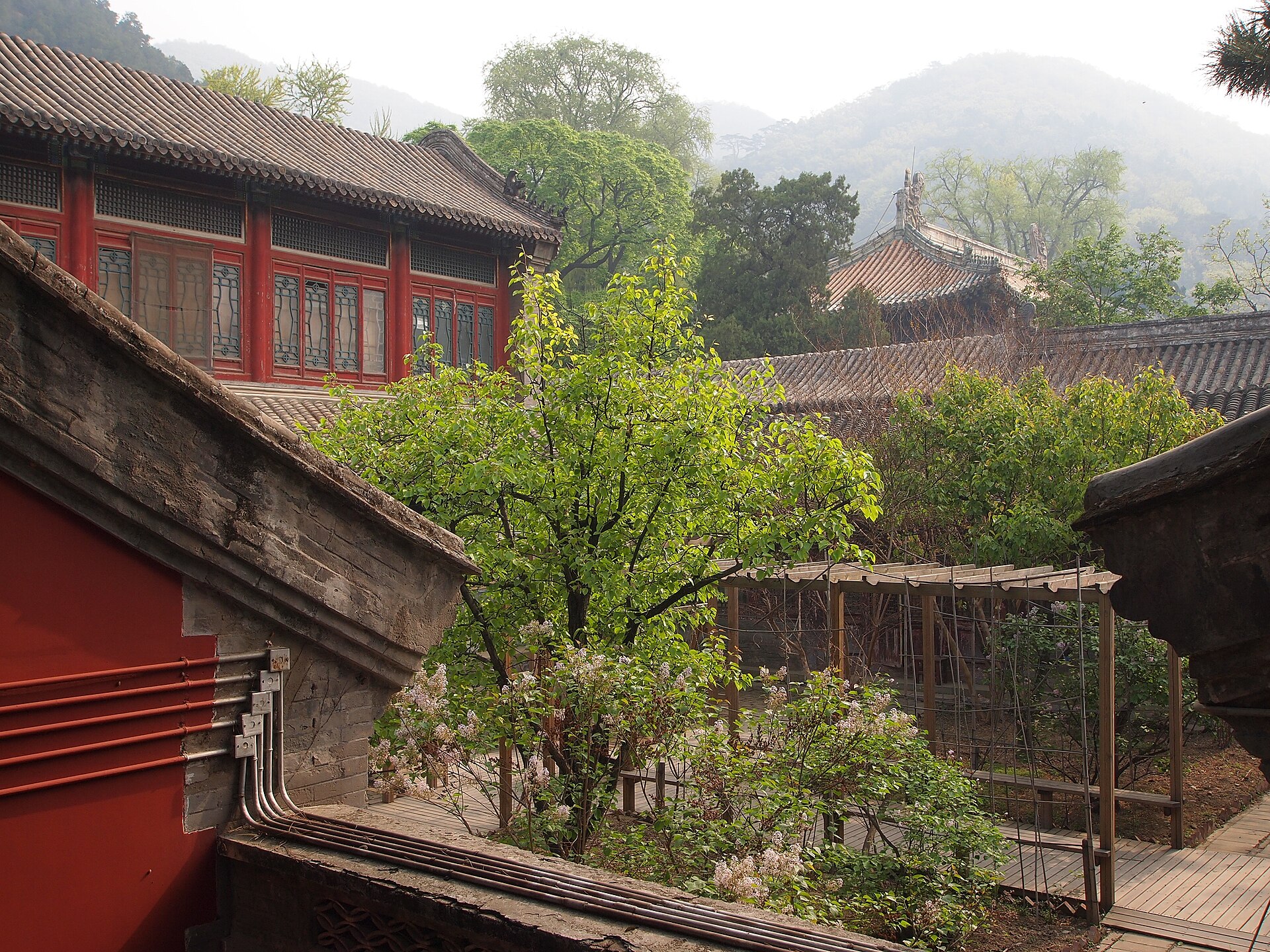 Red halls and tiled roofs surround a leafy courtyard at Tanzhe Temple, with wooded Beijing hills in the background.