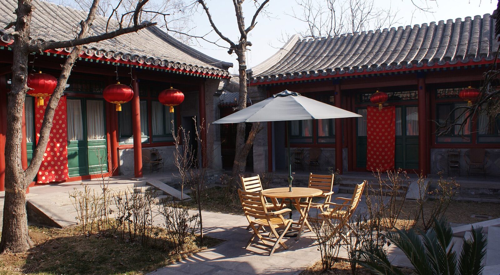Red lanterns hang outside grey-brick halls around a Beijing hotel courtyard with trees, wooden chairs and a parasol.