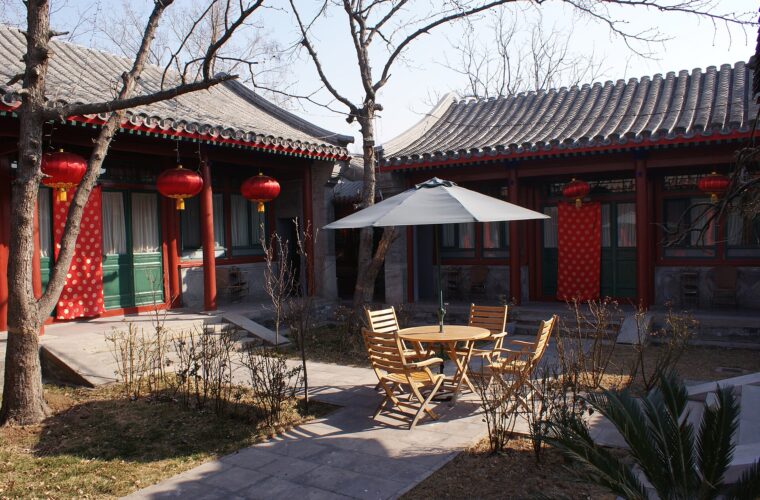 Red lanterns hang outside grey-brick halls around a Beijing hotel courtyard with trees, wooden chairs and a parasol.