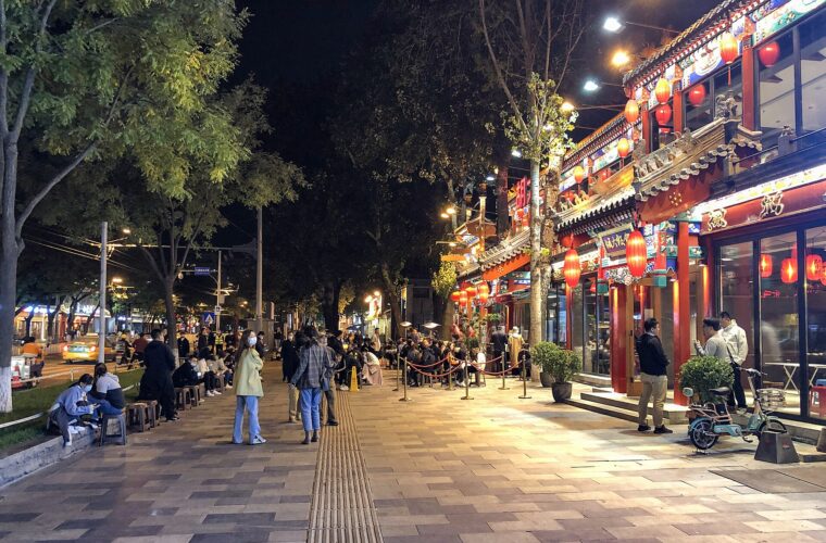 Red lanterns illuminate Guijie restaurants in Beijing as visitors walk and sit along the tree-lined sidewalk at night.
