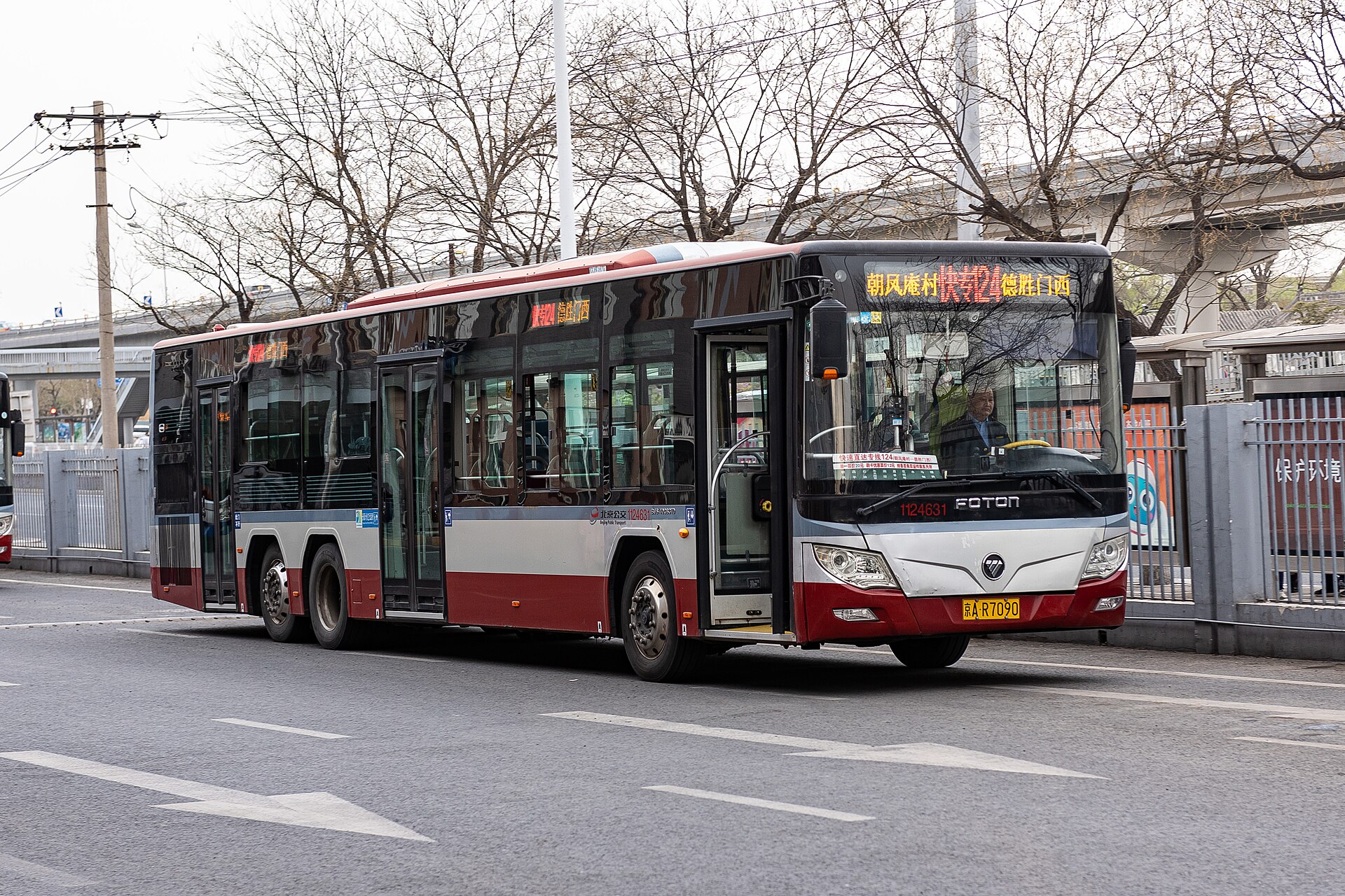 A red and silver Beijing public bus stands at Deshengmenxi beneath bare trees beside an elevated roadway.