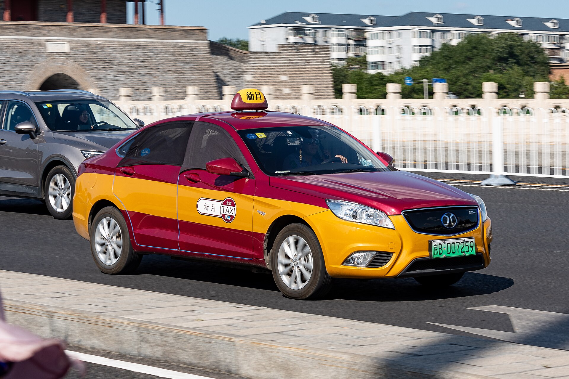Red and yellow Beijing taxi drives along a road at Yongdingmen, carrying a roof sign and taxi door badge.