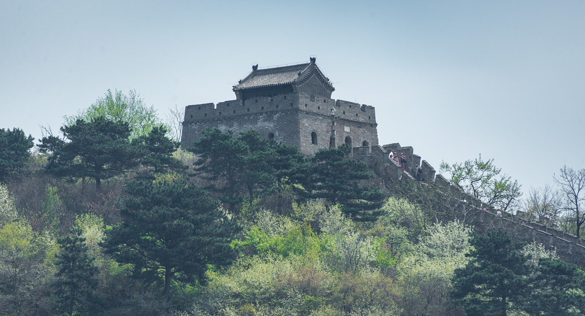 A roofed stone watchtower rises above green pines at Mutianyu, with visitors beside the crenellated wall.