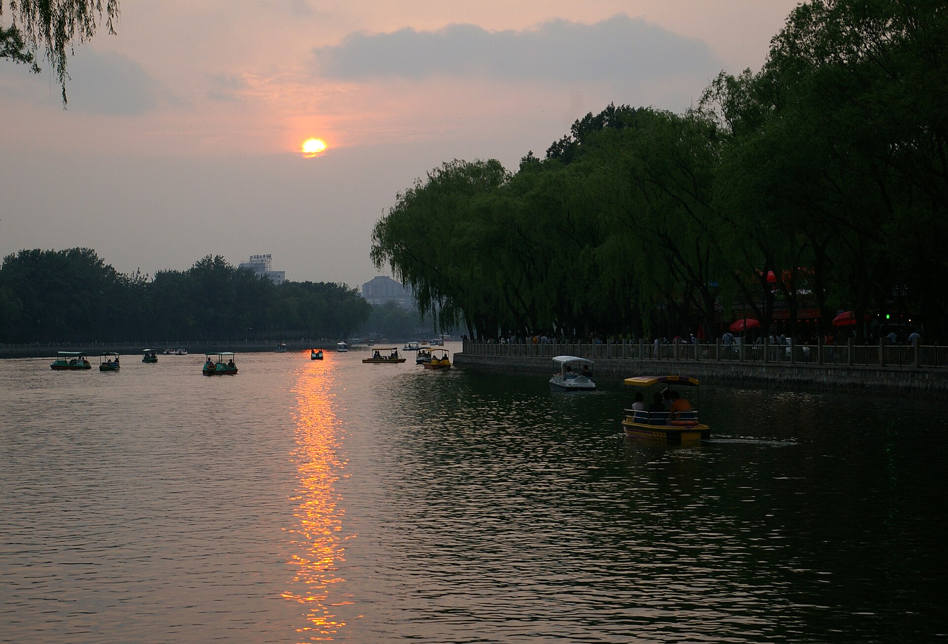 The setting sun casts an orange reflection across Houhai Lake in Beijing, with small boats beside a willow-lined waterfront.