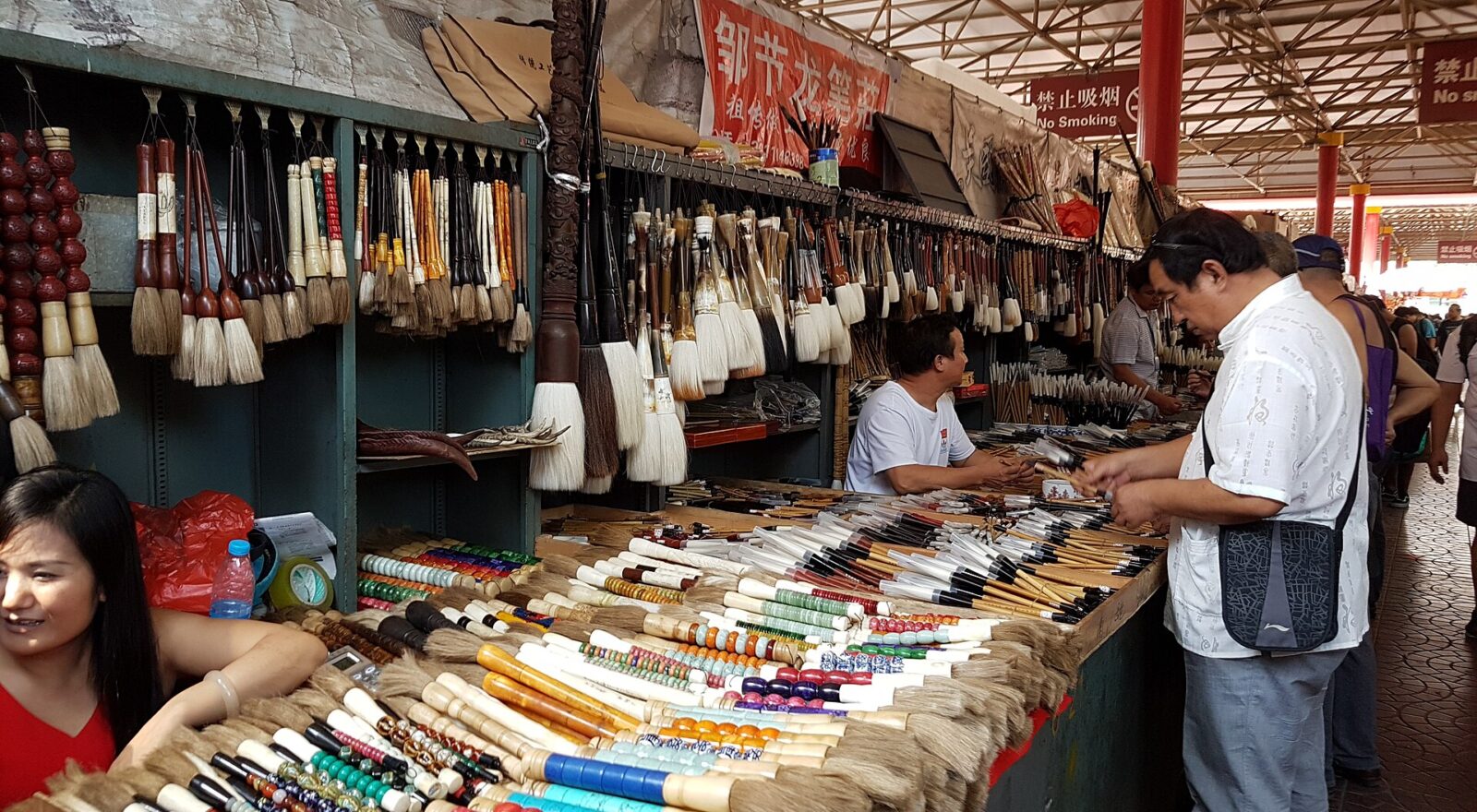 A shopper examines brushes across from a seated vendor at a densely stocked calligraphy stall in Beijing's Panjiayuan market.