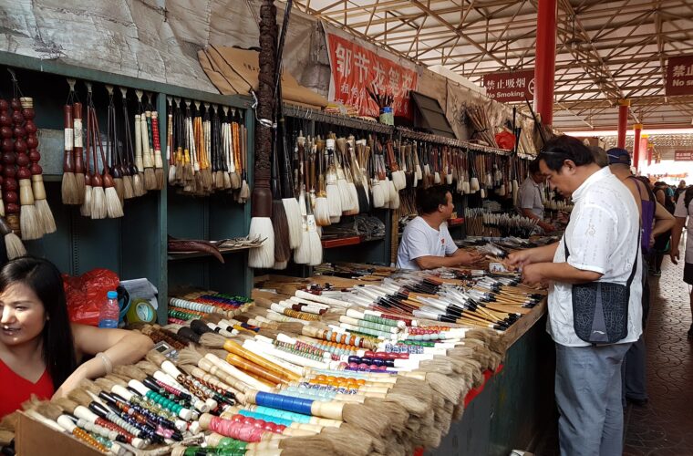 A shopper examines brushes across from a seated vendor at a densely stocked calligraphy stall in Beijing's Panjiayuan market.
