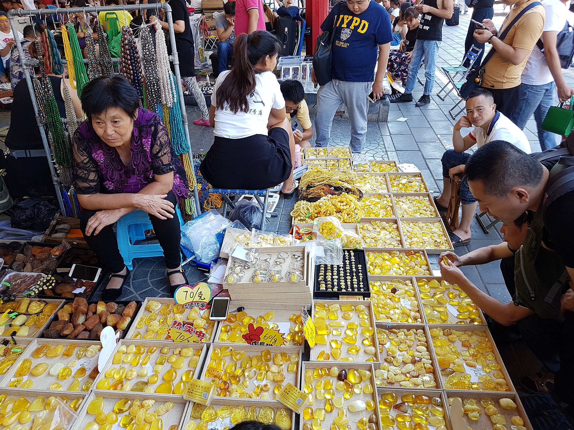 A shopper examines yellow ornaments beside seated stallholders and hanging necklaces at Beijing's busy Panjiayuan market.