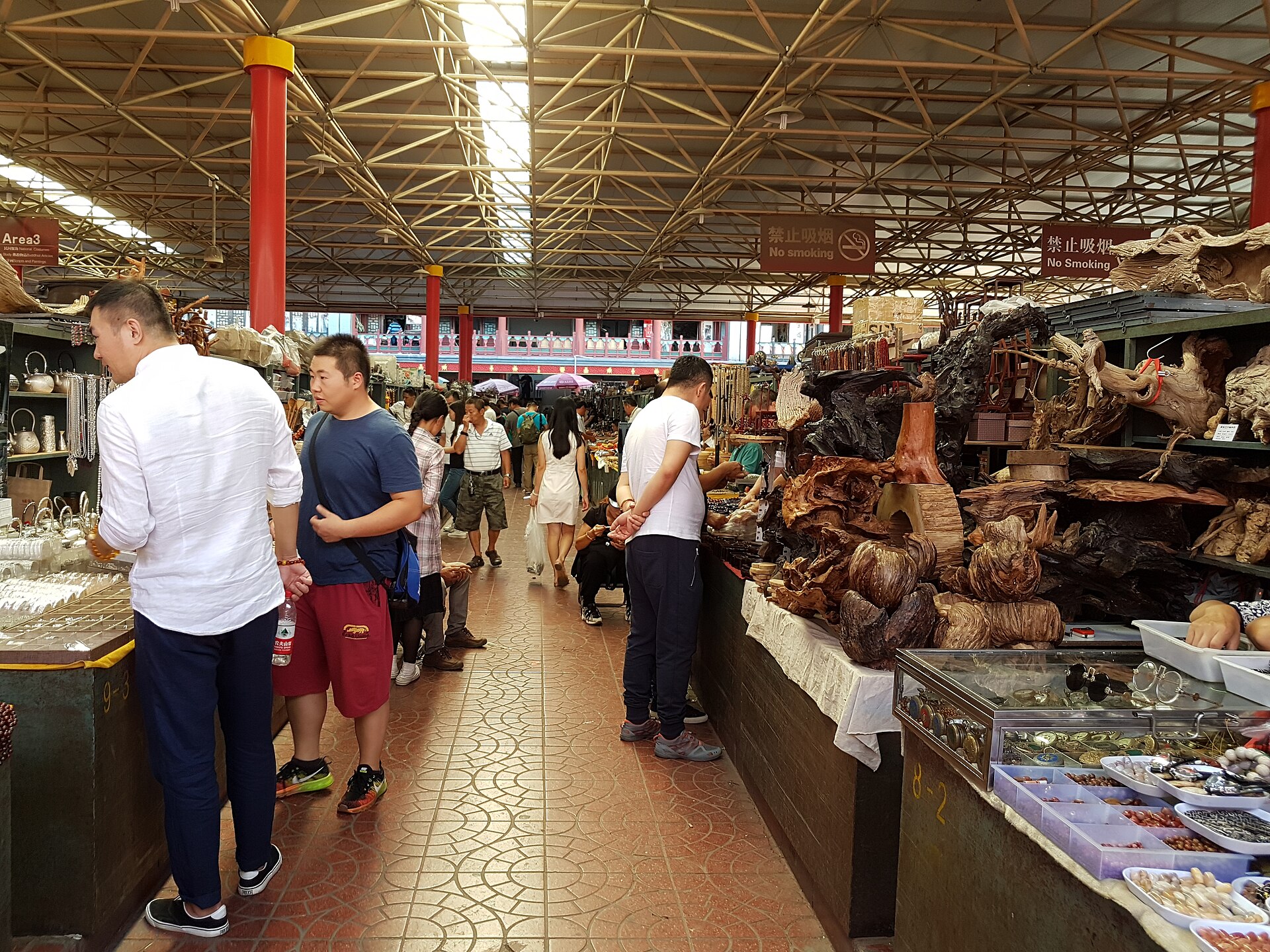 Shoppers and stallholders fill a covered aisle at Beijing's Panjiayuan market, between carved wooden objects and trays of small goods.
