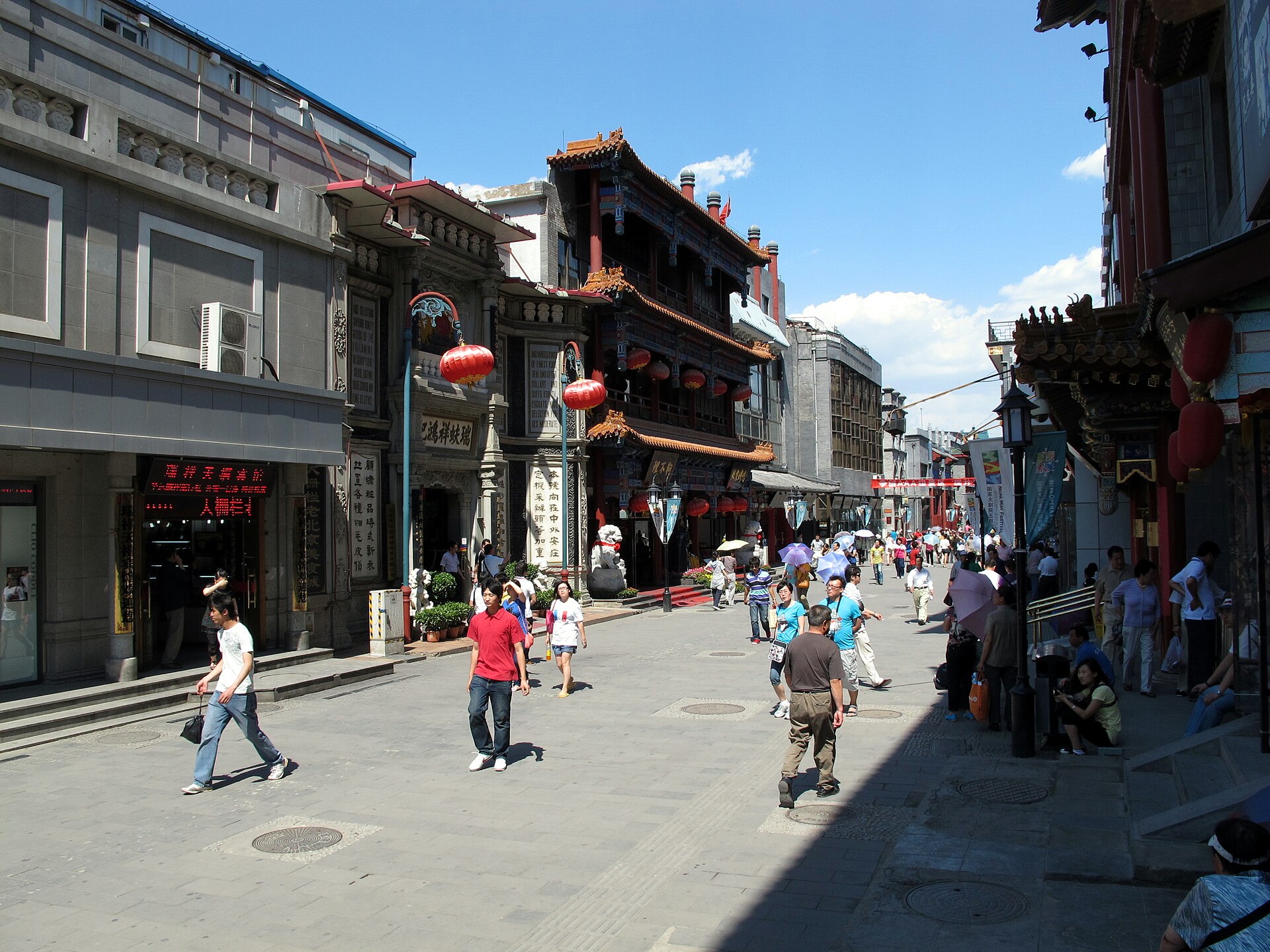 Shoppers walk along Dashilar in Beijing beneath red lanterns beside ornate stone and painted wooden shopfronts.