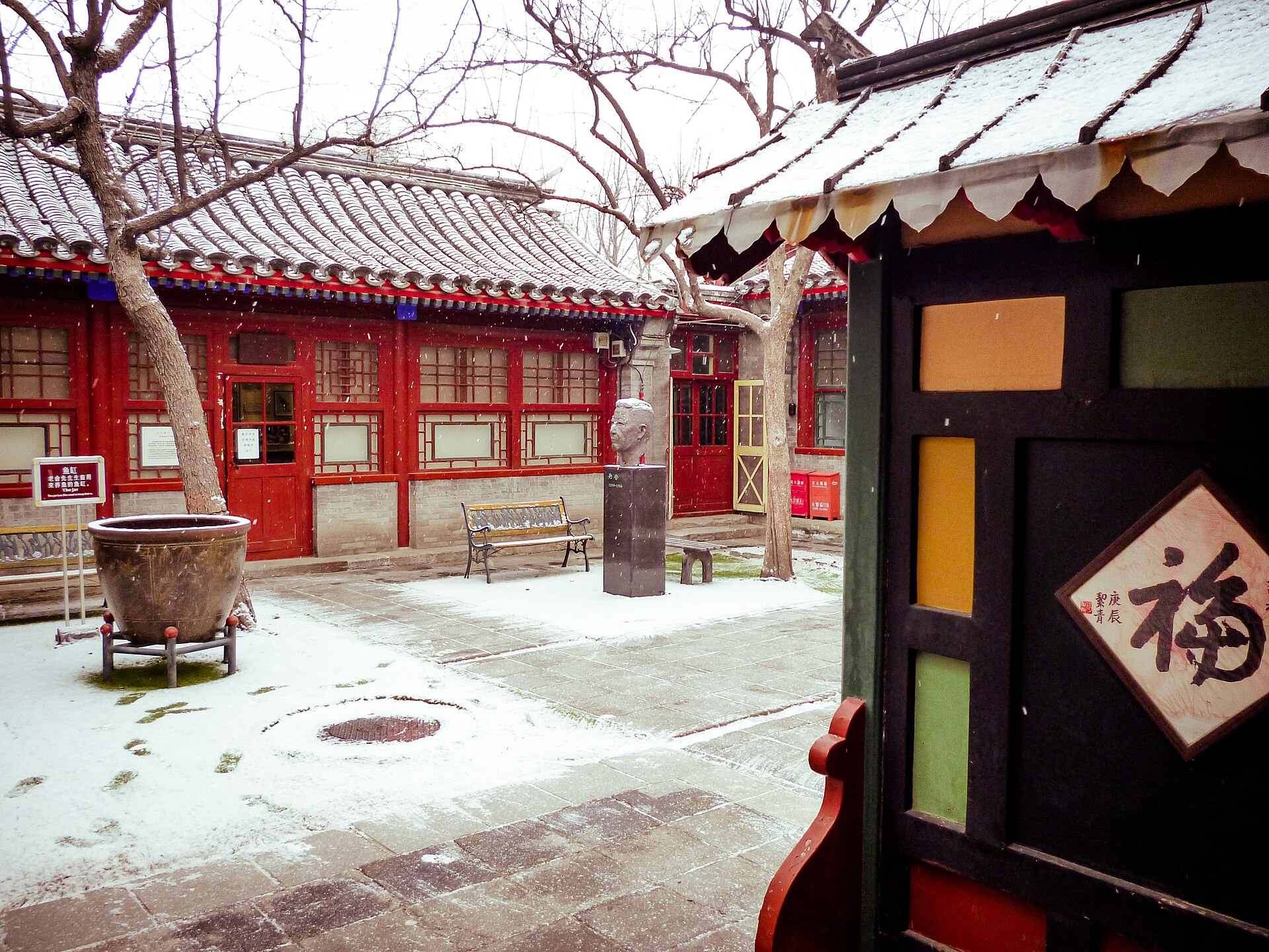 Snow dusts the tree-filled courtyard at Beijing's Lao She Memorial Hall, with red lattice doors and grey tiled roofs.