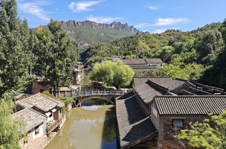 A stone bridge crosses the canal between tiled houses at Gubei Water Town, with wooded mountains rising beyond.