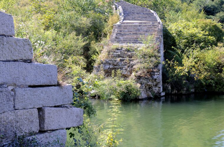 Stone steps on the Great Wall descend into green reservoir water at Huanghuacheng, surrounded by thick foliage and broken masonry.