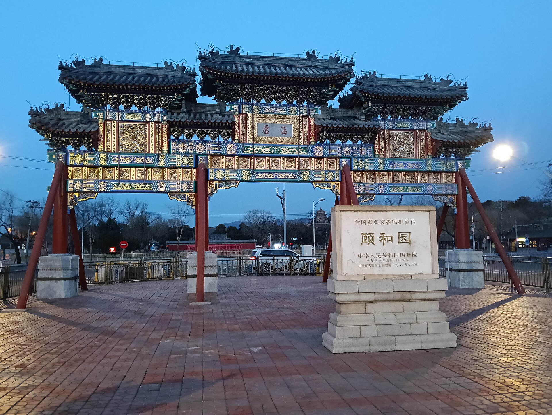An ornate painted memorial archway at dusk outside the East Palace Gate of the Summer Palace, beside a stone heritage tablet inscribed with the palace's Chinese name