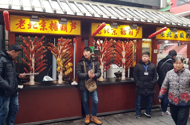 Tanghulu skewers stand in branching displays outside a Wangfujing snack stall in Beijing, with visitors beside the counter.