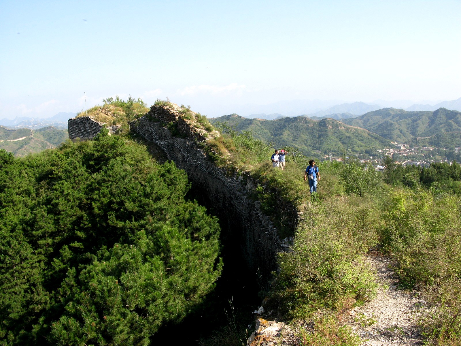 Three hikers walk along an overgrown stretch of the Great Wall at Gubeikou, beside crumbling stone parapets and forested mountain ridges.