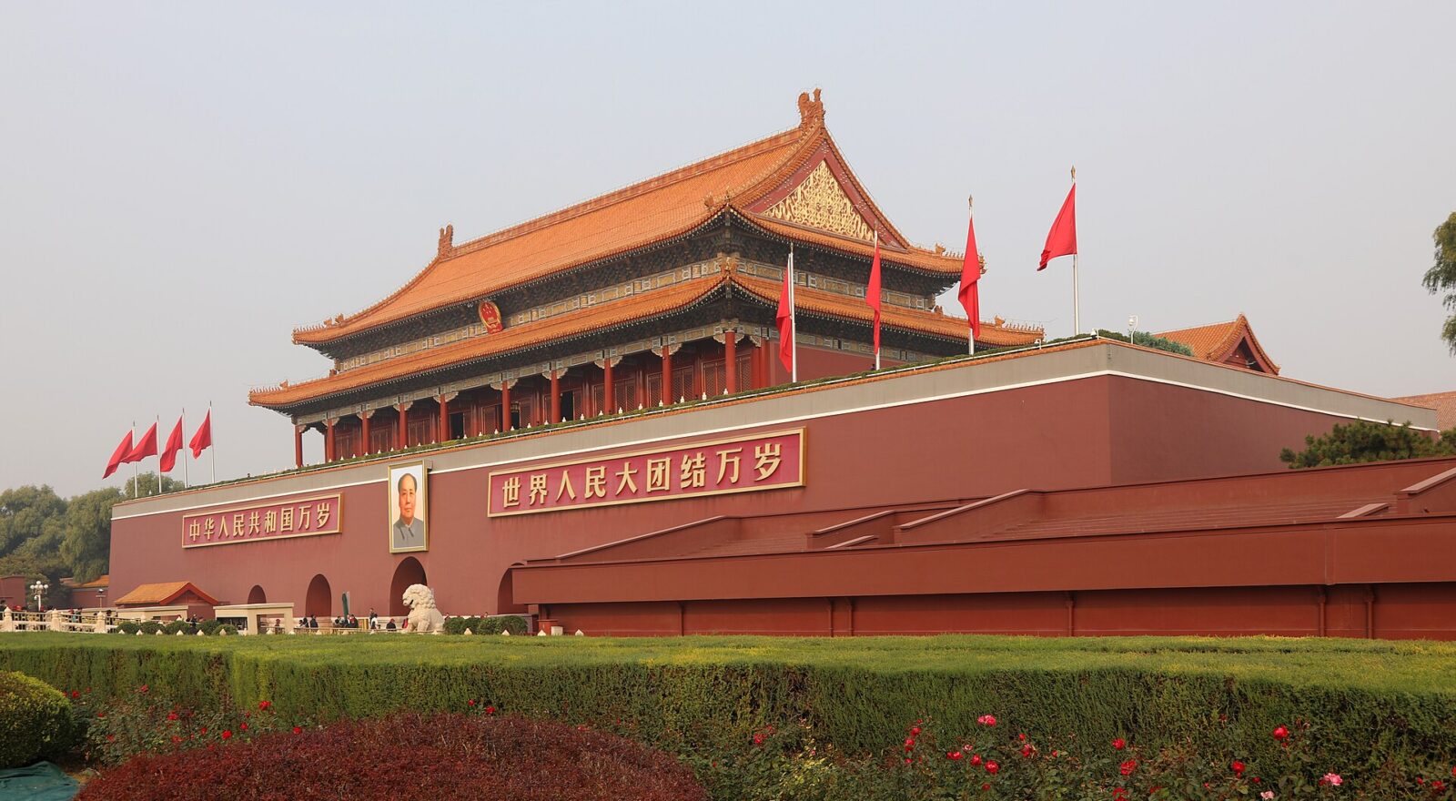 Tiananmen rises above clipped hedges in Beijing, with golden roofs, red flags and small groups of visitors along its base.