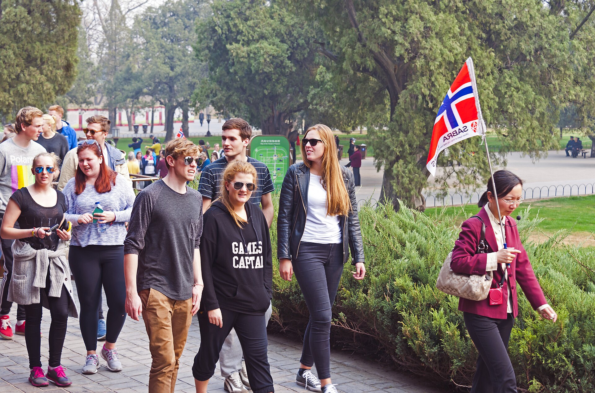 A guide holding a small flag on a pole leads a group of young foreign tourists along a path through the cypress groves of the Temple of Heaven