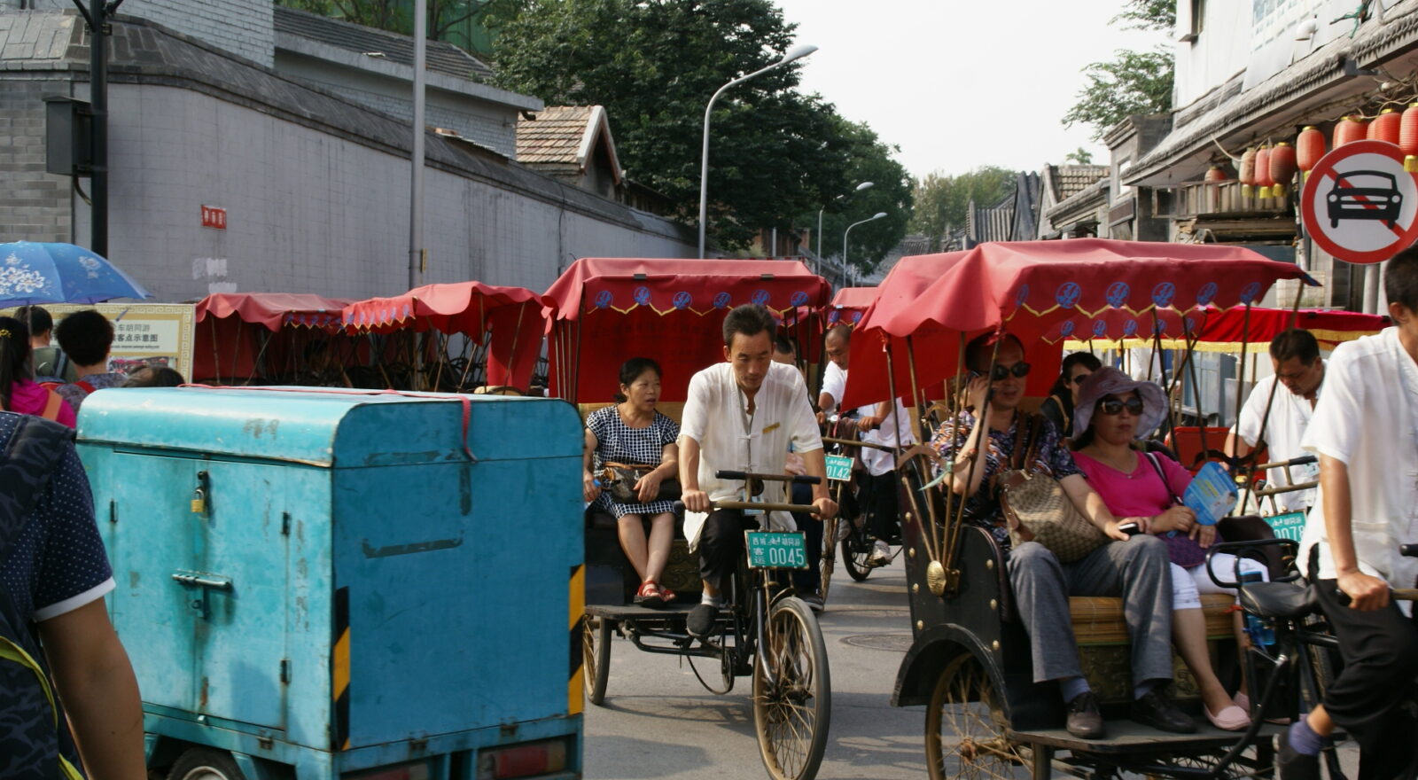 Tourists sit in red-canopied pedicabs as drivers pedal them along Dingfu Jie in Beijing, beside a grey hutong wall and red lanterns.