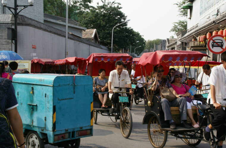 Tourists sit in red-canopied pedicabs as drivers pedal them along Dingfu Jie in Beijing, beside a grey hutong wall and red lanterns.
