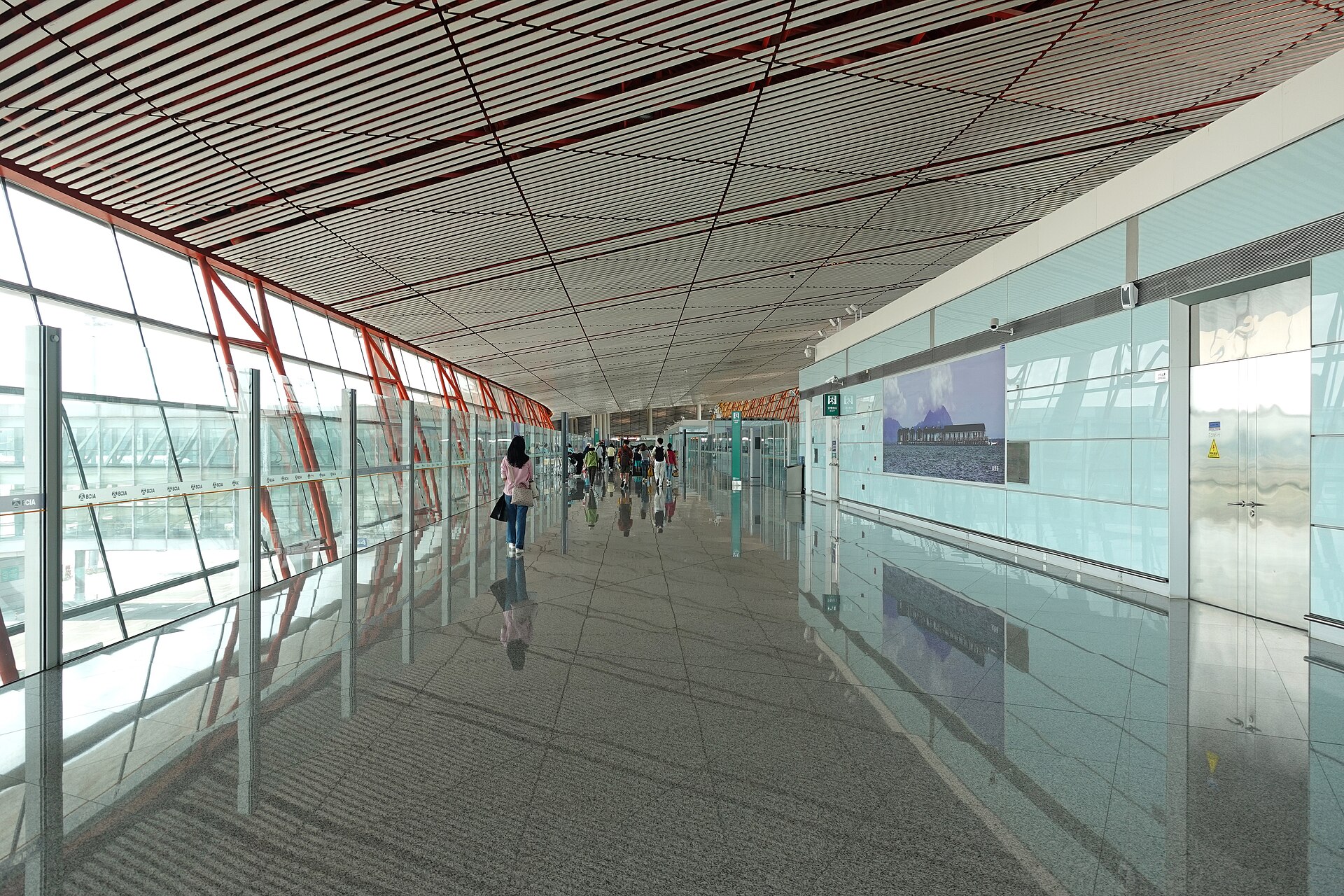 Travellers follow a bright glass-sided international arrivals corridor in Beijing Capital Airport Terminal 3, beneath a ribbed ceiling.